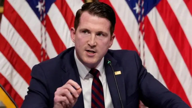 Colin M. McDonald, Umushinjacyaha mukuru wungirije wa Amerika, ariko avugira kuri mikoro, inyuma ye hari idarapo rya Amerika. Yambaye kositimu y